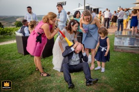Amusing wedding moment captured at Ristorante Erard in Montecarotto, Marche, Italy: man sitting on grass, surrounded by women, holding his ears while woman blasts a horn at his head.